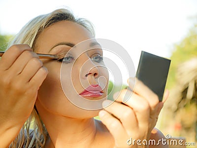 Young beautiful and attractive blond woman with blue eyes retouching makeup with brush applying eyeshadow holding small mirror in Stock Photo