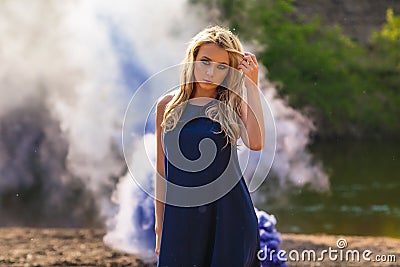 Young beautiful blond woman in blue mini dress and floral wreath standing and trying water on summer day with coutryside Stock Photo