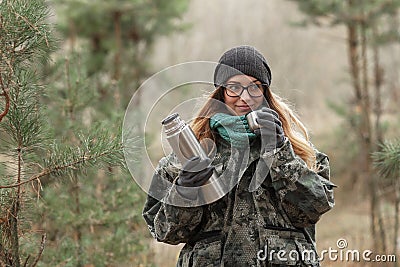 Young beautiful blond woman in camouflage outfit and green scarf posing with thermos in the forest. Travel lifestyle concept. Stock Photo