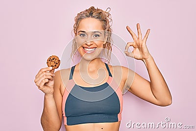 Young beautiful blonde sporty woman doing sport holding cookie over isolated pink background doing ok sign with fingers, smiling Stock Photo