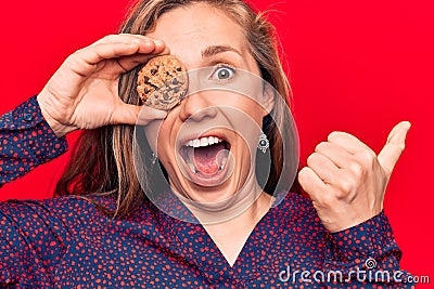 Young beautiful blonde woman holding chocolate chips cookie pointing thumb up to the side smiling happy with open mouth Stock Photo