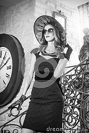 Young beautiful brunette woman in black standing on stairs near an over sized wall clock. Elegant romantic mysterious lady Stock Photo