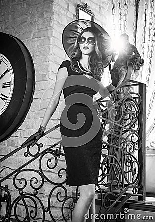 Young beautiful brunette woman in black standing on stairs near an over sized wall clock. Elegant romantic mysterious lady Stock Photo