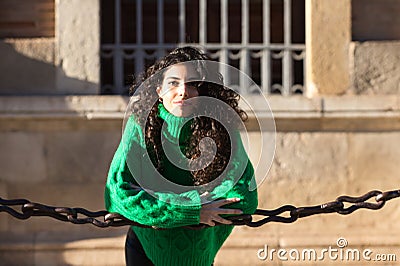 young beautiful brunette woman with curly hair and green woollen coat is leaning on the chains surrounding the cathedral of Stock Photo