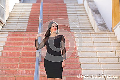 Young, beautiful brunette woman is dressed elegantly in a black dress with transparencies. The girl is walking down a staircase in Stock Photo