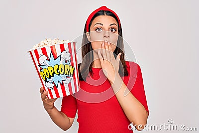 Young beautiful brunette woman eating pack of popcorns snack over isolated white background cover mouth with hand shocked with Stock Photo