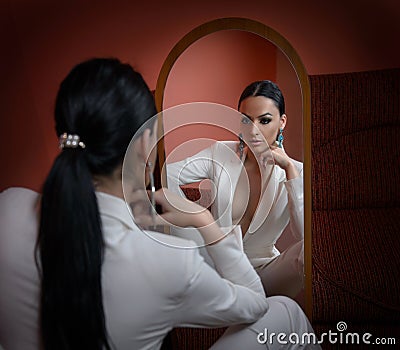 Young beautiful brunette woman in elegant white suit with trousers looking into large mirror. Seductive dark hair girl posing Stock Photo