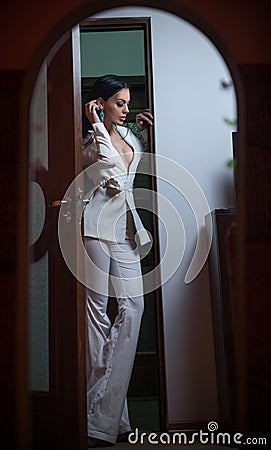 Young beautiful brunette woman in elegant white suit with trousers standing in door frame. Seductive dark hair girl posing indoors Stock Photo