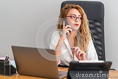 Young beautiful business woman is speaking with phone and sitting confidently on her desk, to check her work. Stock Photo