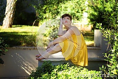 Young beautiful caucasian bored displeased girl with short haircut in short yellow dress sitting outdoors and thinking. sad Stock Photo