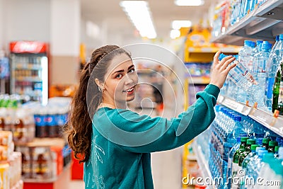 A young beautiful Caucasian woman smiles and takes a bottle of water from the shelf. The concept of shopping and purchasing goods Stock Photo