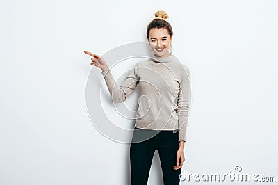 Young beautiful cheerful woman with hair bun wearing in jeans and sweater pointing with forefinger on copy space over white wall Stock Photo