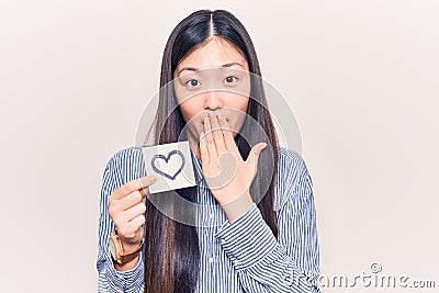 Young beautiful chinese woman holding reminder with heart shape covering mouth with hand, shocked and afraid for mistake Stock Photo