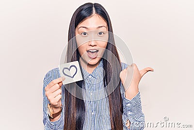 Young beautiful chinese woman holding reminder with heart shape pointing thumb up to the side smiling happy with open mouth Stock Photo