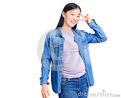 Young beautiful chinese woman pregnant expecting baby doing peace symbol with fingers over face, smiling cheerful showing victory Editorial Stock Photo
