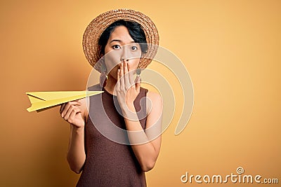 Young beautiful chinese woman on vacation wearing summer hat holding paper airplane cover mouth with hand shocked with shame for Stock Photo