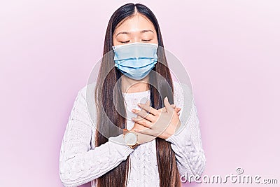 Young beautiful chinese woman wearing medical mask smiling with hands on chest, eyes closed with grateful gesture on face Editorial Stock Photo