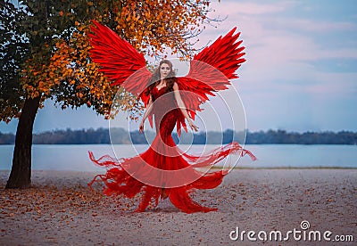 Young beautiful fantasy woman fallen angel lying in air near a tree with orange leaves. Creative red costume, huge Stock Photo