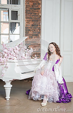 Young beautiful flirtatious woman in fantasy rococo style medieval dress sitting near piano with pink flowers Stock Photo