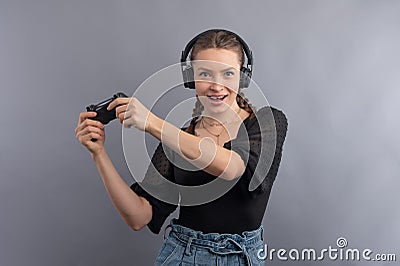 Young beautiful gamer girl playing video game using joystick and headphones on grey background. Girl in casual wear with emotional Stock Photo