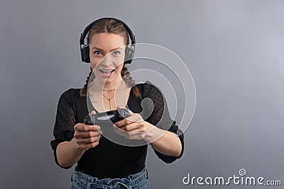 Young beautiful gamer woman playing video game using joystick and headphones on grey background. Woman in casual wear with Stock Photo