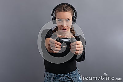 Young beautiful gamer woman playing video game and using joystick and headphones on grey background. Emotional face. Gambler Stock Photo