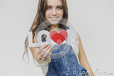 A young beautiful girl is on a gray background. Dressed in a denim suit and a white T-shirt. Holds a sign with a word of Stock Photo