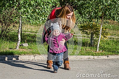 Young beautiful girl with long hair in a jacket mother teaches his daughter to walk their baby's first steps outdoors on a sunny d Stock Photo