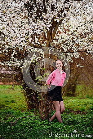 Young beautiful girl in a pink shirt standing under blossoming apple tree and enjoying a sunny day. Stock Photo