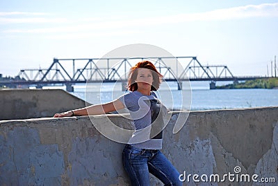 The young beautiful girl with red hair poses against the background of the river and the railway bridge Stock Photo