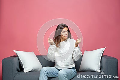 Young beautiful girl show money gesture with fingers, asking for debt, salary payment or fee sitting on sofa Stock Photo