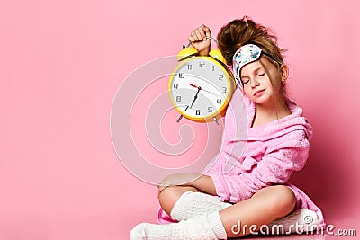 Yawning teenage girl holding an alarm clock while sitting in a housecoat on the floor. Stock Photo