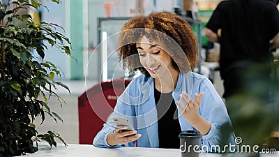 Young beautiful girl teenager millennial African American woman with curly hair sits in cafe looking at mobile phone Stock Photo