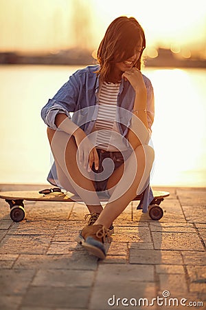 A young beautiful girl thoughtfully sitting on a skateboard at the dock and enjoying the sunset on the sea. Summer, sea, vacation Stock Photo