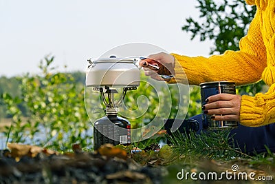 Young beautiful girl in a yellow sweater makes coffee in the forest on a gas burner. Making coffee on a primus stove in the autumn Stock Photo