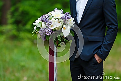 Young beautiful groom in dark blue suit standing and holding big wedding bouquet with lilac and long ribbons. Spring, park outdoor Stock Photo