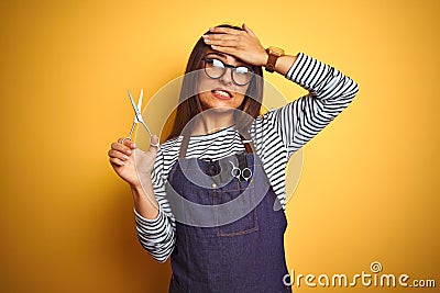 Young beautiful hairdresser woman holding scissors standing over isolated yellow background stressed with hand on head, shocked Stock Photo