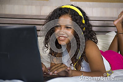 Young beautiful happy black hispanic woman at home bedroom lying cheerful on bed listening to internet music with headphones and l Stock Photo