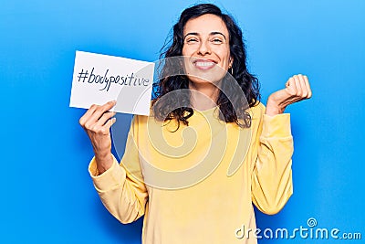 Young beautiful hispanic woman holding paper with hashtag body positive screaming proud, celebrating victory and success very Stock Photo