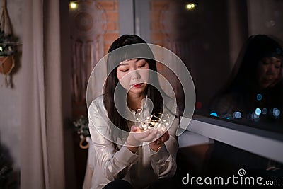 A young, beautiful Korean Asian woman holds a lighted festive garland in her cozy house with dreams of positive emotions Stock Photo