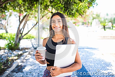 Young beautiful latin student with laptop and cup of coffee studying in the park. Girl is walking in the park with great smile and Stock Photo