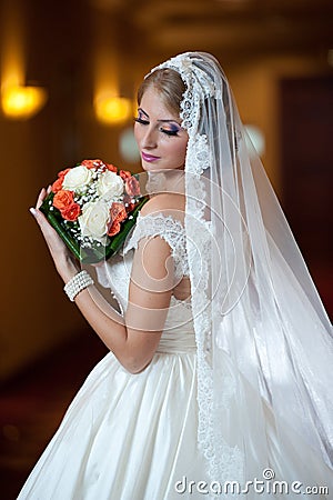 Young beautiful luxurious woman in wedding dress posing in luxurious interior. Bride with long veil holding her wedding bouquet Stock Photo