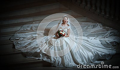 Young beautiful luxurious woman in wedding dress sitting on stair steps in semi-darkness. Bride with huge wedding dress Stock Photo