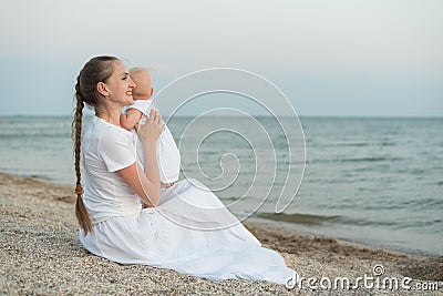 Young beautiful mom sitting on beach and hugging baby. Mothers love and care Stock Photo