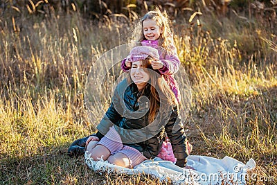 Young beautiful mother with her daughter on a walk on a sunny autumn day. Daughter is trying to put her hat on mother, they are la Stock Photo