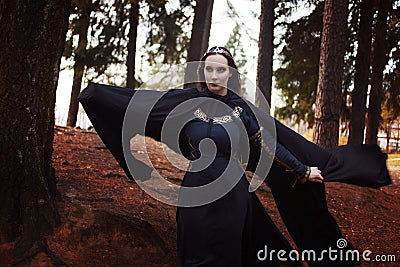 Young beautiful and mysterious woman in woods, in black cloak with hood, image of forest elf or witch Stock Photo