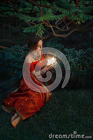 Young beautiful oriental indian fantasy woman sits on green grass, background night magic forest. Red vintage saree Stock Photo