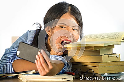 Young beautiful and playful Asian Korean student teenager taking selfie picture on desk with mobile phone biting pile of books in Stock Photo