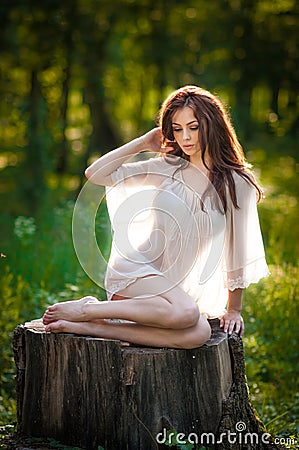 Young beautiful red hair woman wearing a transparent white blouse posing on a stump in a green forest. Fashionable girl Stock Photo