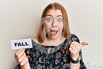 Young beautiful redhead woman holding fall word on paper pointing thumb up to the side smiling happy with open mouth Stock Photo